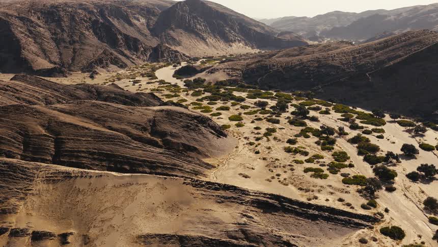 This aerial view captures a winding riverbed nestled between rugged layered mountains in a desert landscapesmall green trees and shrubs are scattered along the sandy valley floor contrasting with the stark rocky terrain