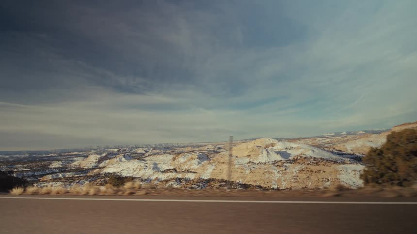 A roadside view captures a vast desert landscape featuring snowdusted plateaus and dry vegetation under a pale blue skythe camera pans steadily from a moving vehicle revealing rolling hills and scattered evergreen trees alongside a paved highway