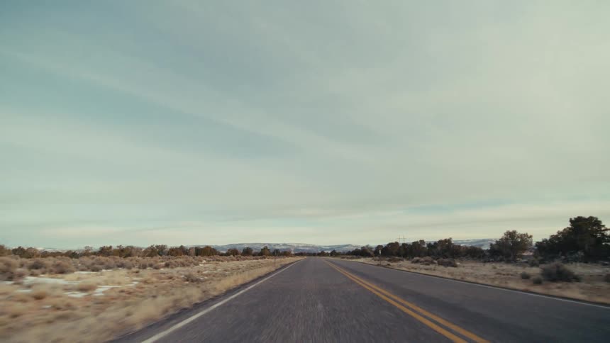 A two lane highway stretching through a vast desert toward distant mountains under a pale blue sky