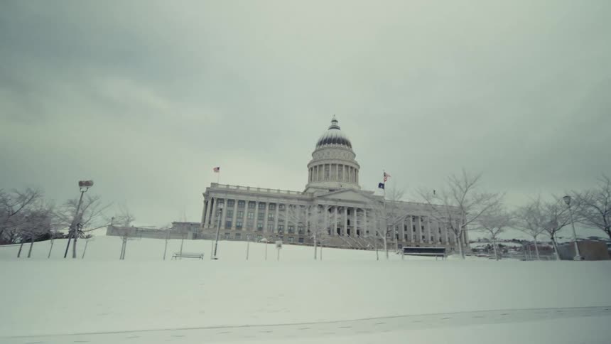 A view from a moving vehicle shows a large stone capitol building with a dome situated on a snowy hill behind bare trees under a grey sky