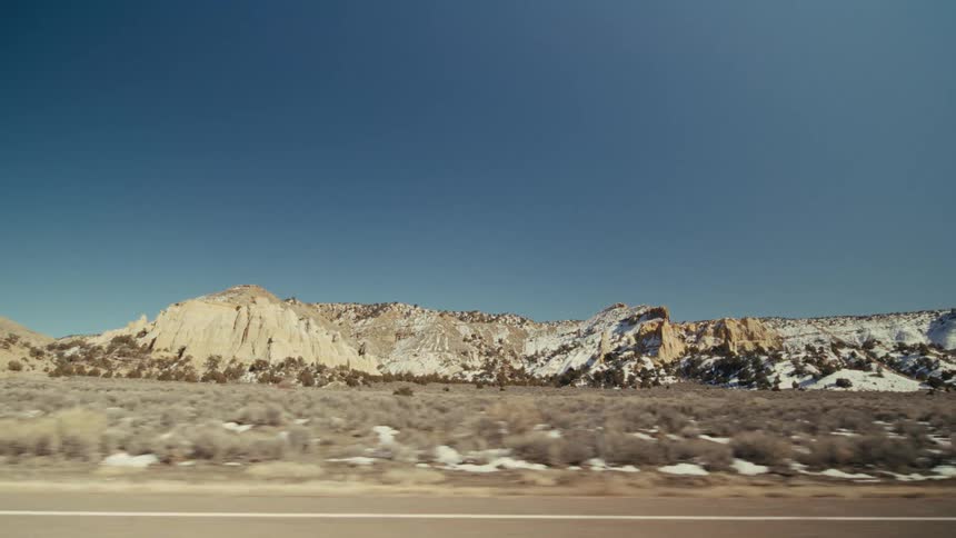 The camera travels alongside a vast desert landscape featuring lightcolored rocky mesas and patches of snow under a clear blue sky lowlying shrubs cover the foreground as the road stretches past the distant rugged mountain formations