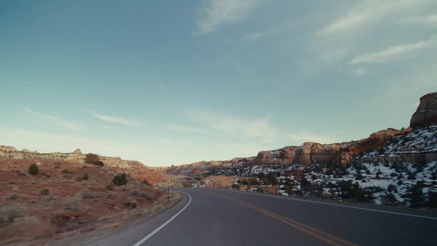 A car travels along a winding twolane highway that cuts through a vast desert canyon landscapethe surrounding terrain features red rock formations and sparse vegetation with patches of snow visible on the distant hillsides under a clear blue sky