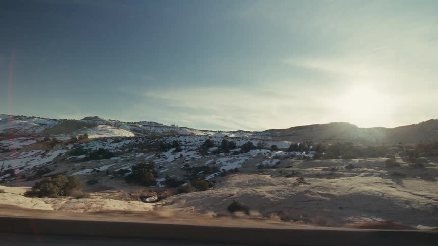 A view from a moving vehicle shows a vast desert landscape with patches of white snow on rocky hills under a bright blue sky with thin clouds