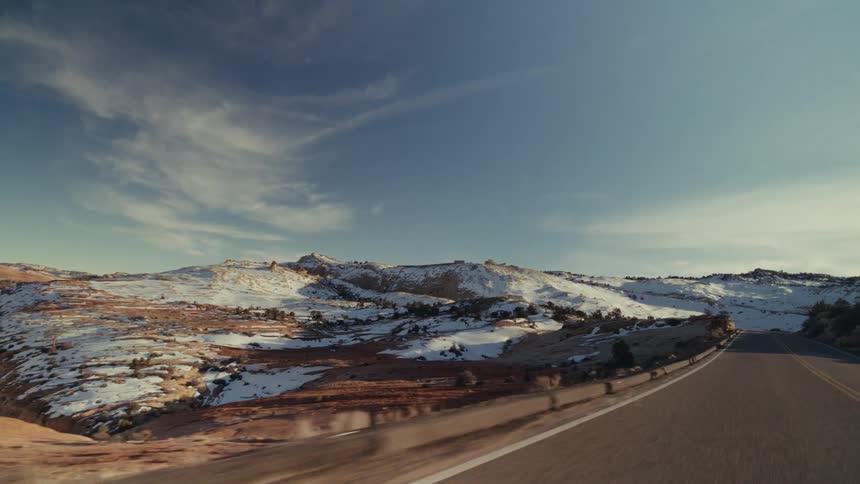 A view from a moving vehicle shows a paved road winding through a vast desert valley with snow covered hills and rocky mountains under a blue sky with soft white clouds