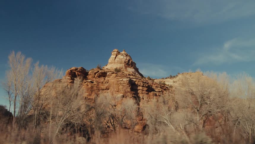 A view from a moving vehicle shows a vast orange desert landscape with large rock formations and dry brush under a clear blue sky