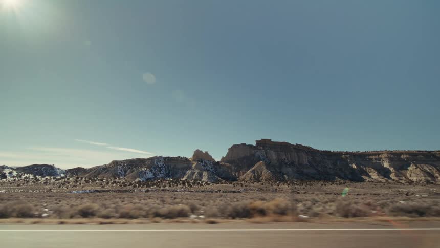 The video shows a sideview perspective from a moving vehicle traveling along a highway through a vast desert landscape large rugged rock formations and mesas sit under a clear blue sky with the bright sun creating a slight lens flare in the upper left