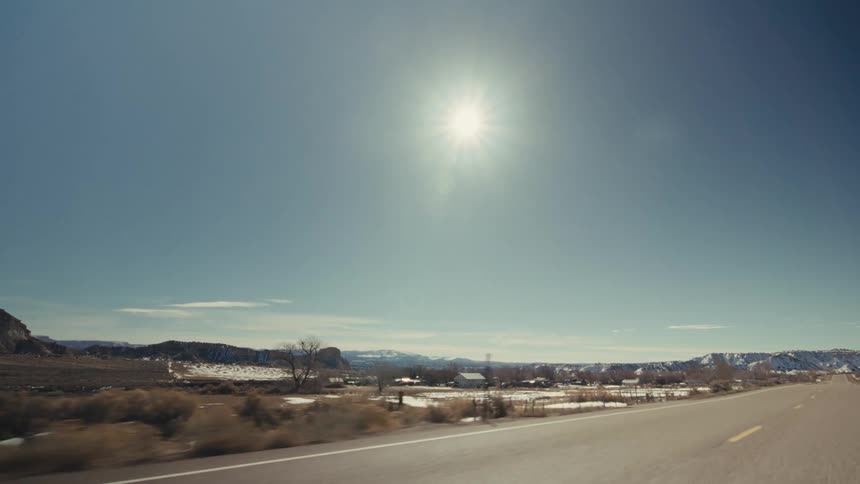 A bright sun hangs high in a clear blue sky over a vast arid landscape as the camera travels along a paved highwaypatches of snow dust the desert floor alongside the road with distant mesas and small buildings visible beneath the intense sunlight