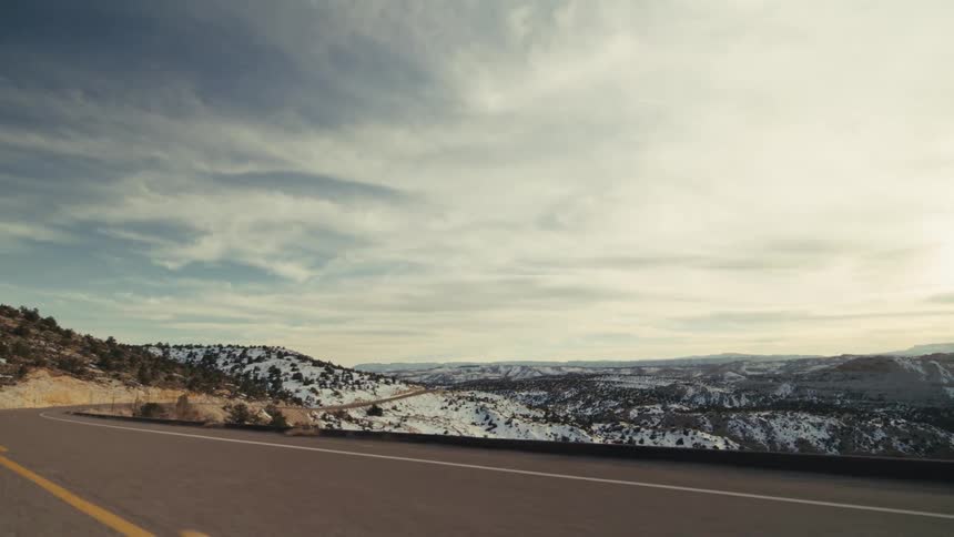 Videoa view from a moving vehicle shows a long paved road winding through a vast valley with snow covered hills and rocky mountains under a bright cloudy sky