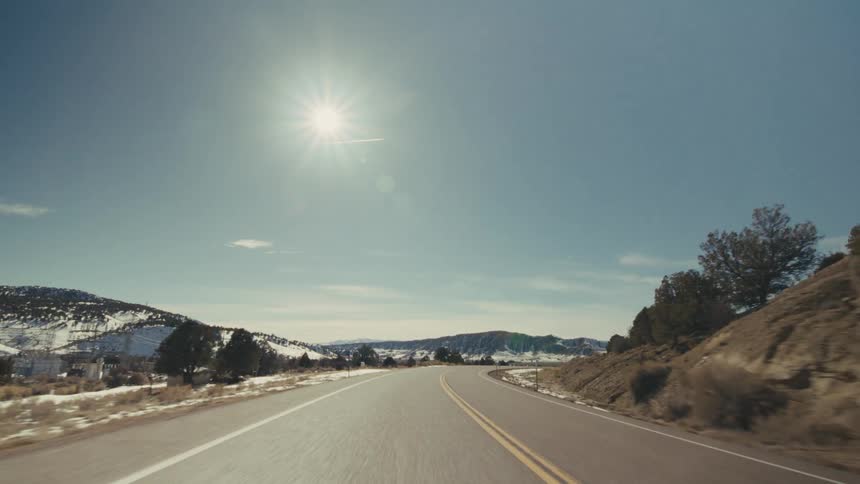 A view from a moving vehicle travels along a straight paved highway with a yellow line through a vast desert valley featuring large rocky mountains and dry vegetation under a bright sun and clear sky