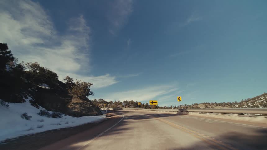 A winding two lane asphalt highway through a mountainous canyon with melting snow on the embankments under a bright blue sky with wispy clouds and scattered evergreen trees