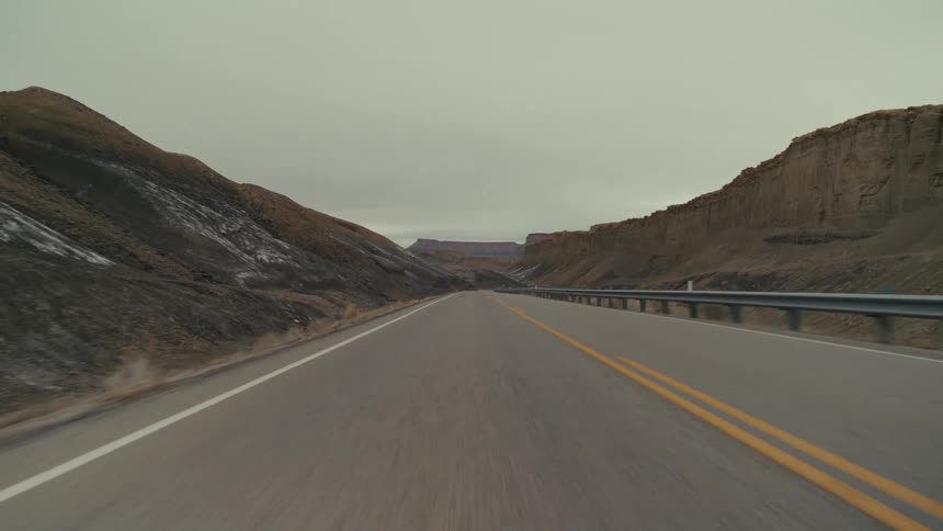 A paved highway winding through a desolate valley of dark rocky mountains and hills under a thick overcast sky