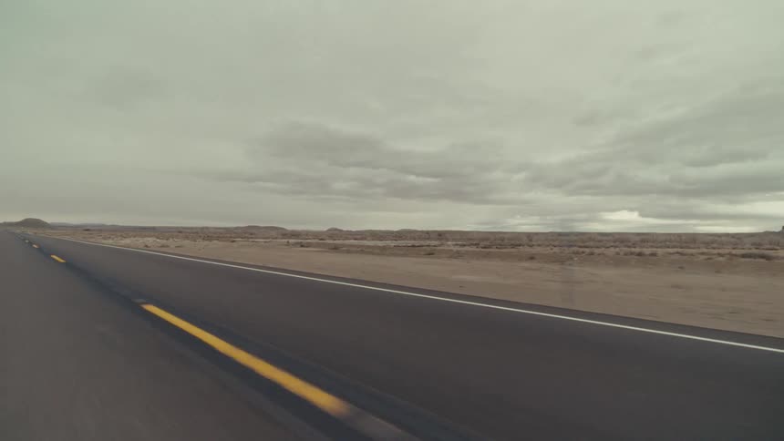A vehicle travels along a paved highway through a desert landscape under an overcast sky
