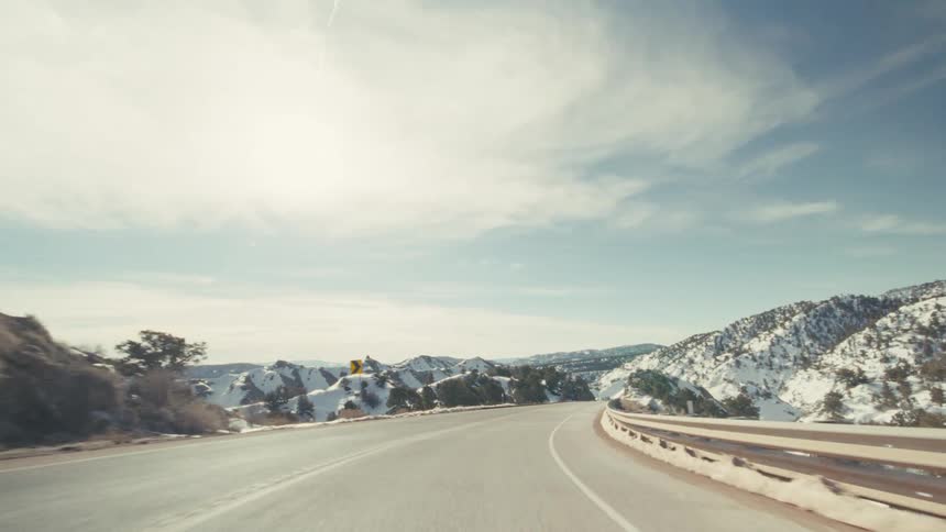 A curved asphalt highway through a vast winter canyon featuring steep rocky hillsides covered in patches of white snow under a bright blue sky with thin clouds and a distant mountain range on the horizon