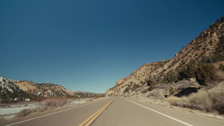 A view from a moving vehicle travels along a curved paved highway with two yellow lines through a rocky canyon with patches of white snow under a blue sky with soft clouds