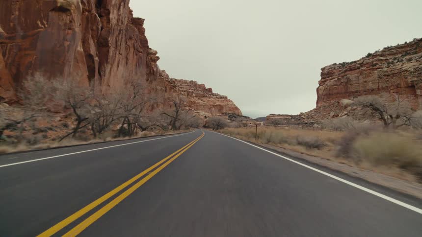 A view from a moving vehicle travels along a curved paved highway with two yellow lines through a rocky canyon under a grey overcast sky