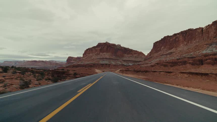 A winding two lane asphalt highway through a vast desert canyon surrounded by massive red rock formations and dry sandy hills under a thick layer of grey clouds