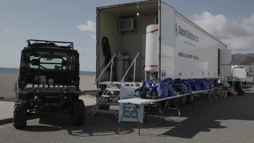 Mobile laundry unit parked near the sea with access stairs utility vehicle beside it and tables lined with bags