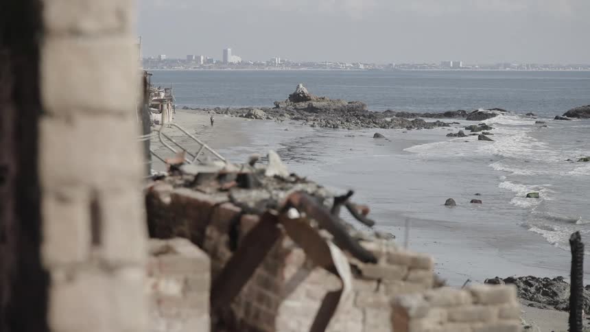 Waves roll over rocks and into shore as a person walks on the beach and buildings across the water