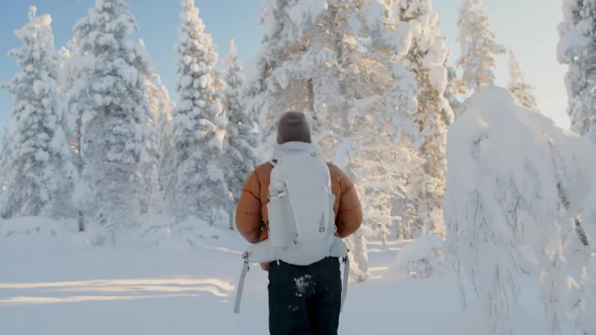 A person wearing an orange jacket and a white backpack walks through deep snow in a winter forest