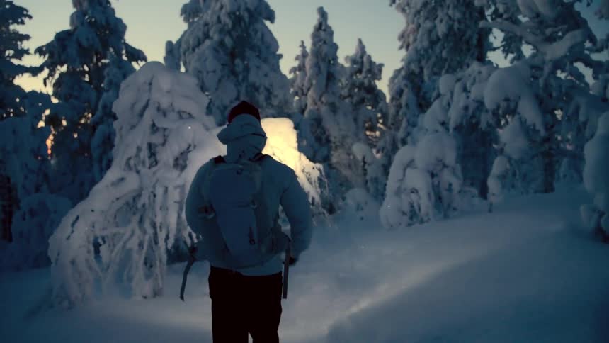 A person with a backpack walks through a snow covered forest at dusk while using a flashlight