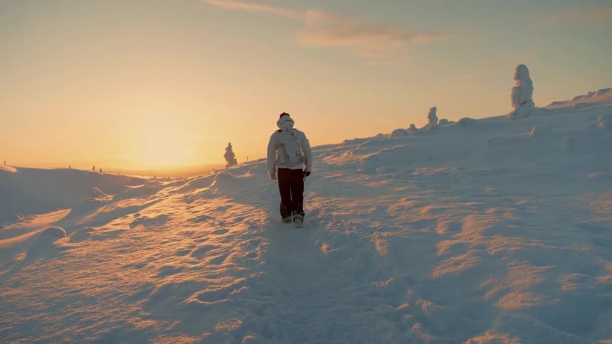 A person wearing a white jacket and a backpack walks away from the camera on a snowy path toward a bright golden sunset in a vast winter landscape