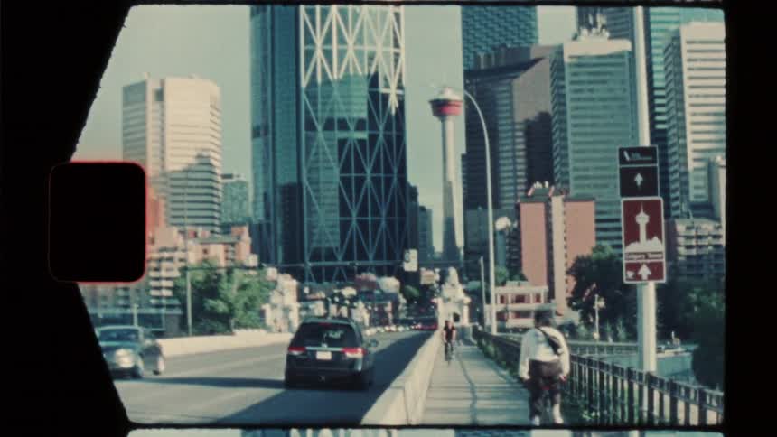 A cyclist and pedestrians travel across a bridge toward a city skyline where several buildings and glass skyscrapers rise under a clear sky while cars drive along the sunlit urban roadway