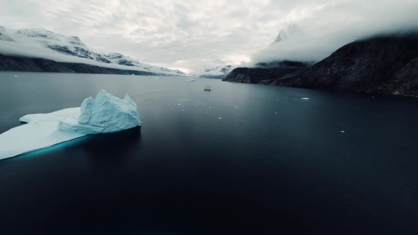This video features a dramatic fpv drone shot that glides through a narrow crevice between two towering ice walls it then opens up to reveal a vast cold bay filled with icebergs and a distant ship under a moody overcast sky