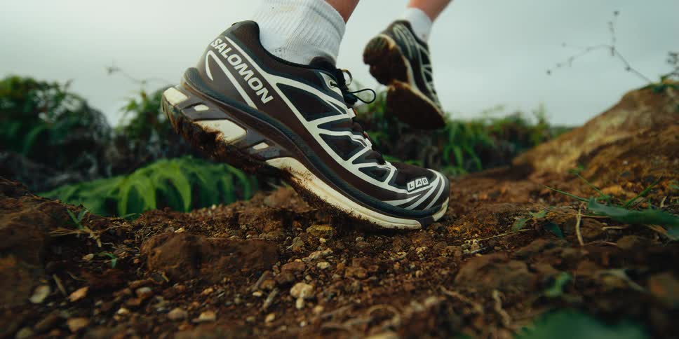 A closeup shot captures a runners salomon shoes kicking up dirt as they sprint along a rugged trail the trail is surrounded by lush green ferns and rolling hills under a cloudy sky
