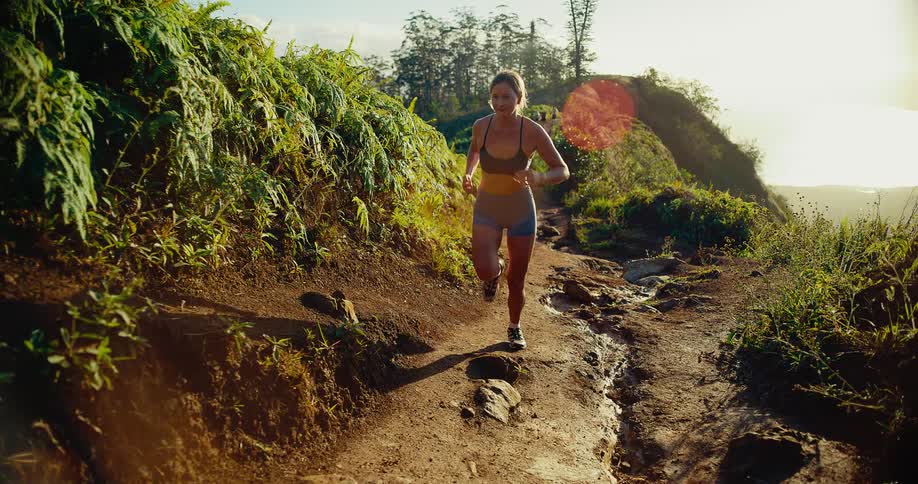 A woman runs toward the camera on a narrow dirt trail along a steep mountain ridge