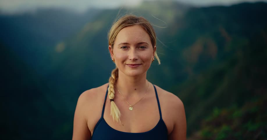 A young woman with a blonde braid and a blue tank top smiles at the camera while standing on a high ridge overlooking a vast green mountain valley under a soft overcast sky