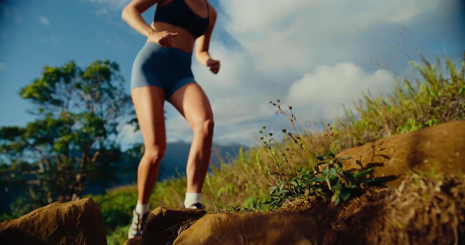 A woman wearing a dark sports bra and blue shorts runs up a steep dirt path under a bright sky