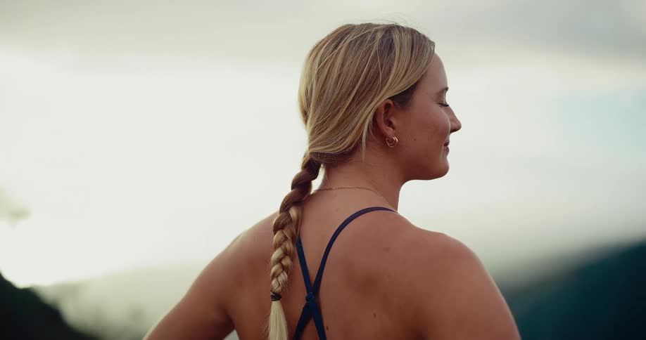 A blonde woman with a long braid stands on a grassy ridge looking out over a vast mountain valley she wears blue athletic gear and keeps her hands on her hips while admiring the scenic view at sunset