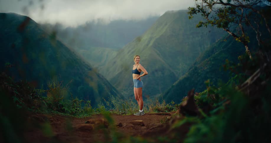 Woman stands on a trail at the edge of a mountain with her hands on her hips as she looks around