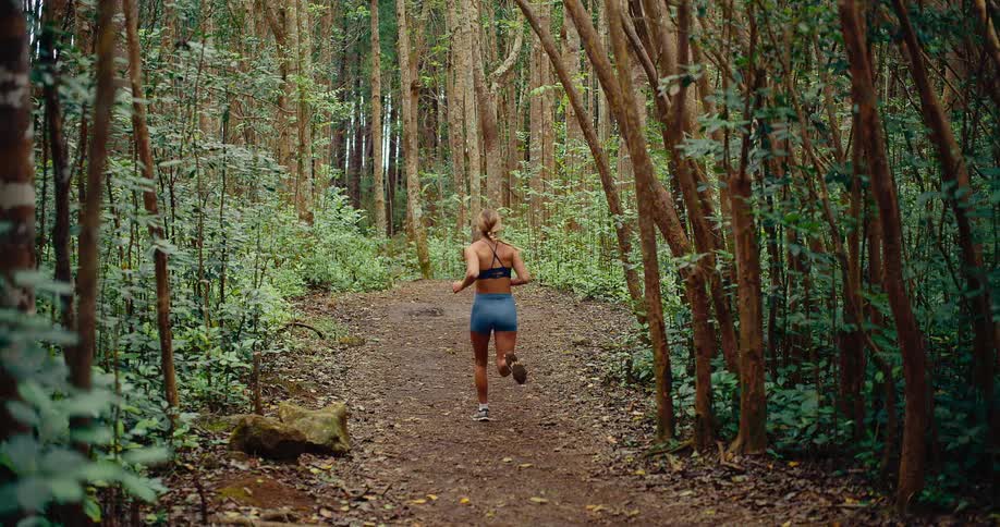 A woman with blonde hair in a braid wearing a blue sports bra and shorts runs away from the camera down a dirt path through a dense forest with many tall thin trees