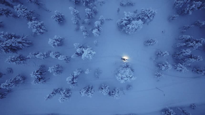 A top view shows a snowmobile with a bright headlight moving slowly along a narrow trail through a dense forest of snow covered trees during the blue hour