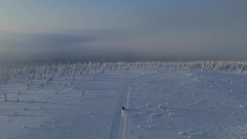 Person rides a vehicle through snowy terrain while heading towards frost covered forest on a winter day