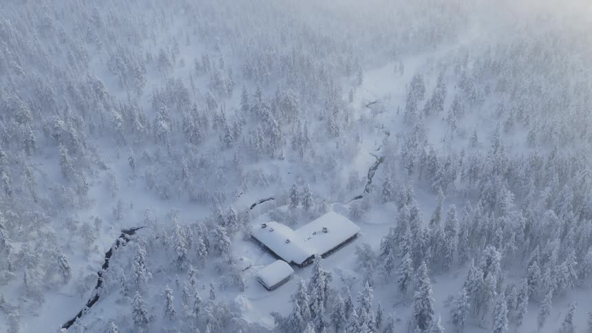 The video shows an aerial drone shot of a snowcovered building nestled deep within a dense frostladen forest a dark winding stream cuts through the white landscape as the camera slowly pulls back to reveal the scale of the wintry wilderness