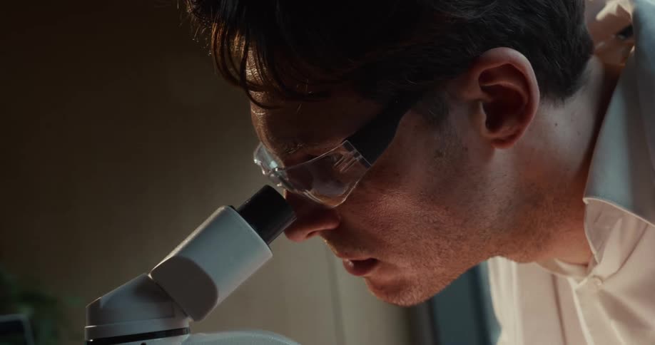 A scientist wearing protective safety glasses peers intensely into the eyepiece of a microscopehis focused expression suggests he is carefully examining a specimen during a detailed laboratory investigation