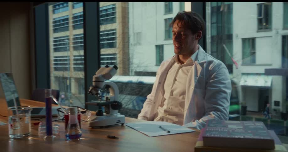 Doctor sits at table looking in a microscope and lifts his head with a smile on his face and writes notes on paper