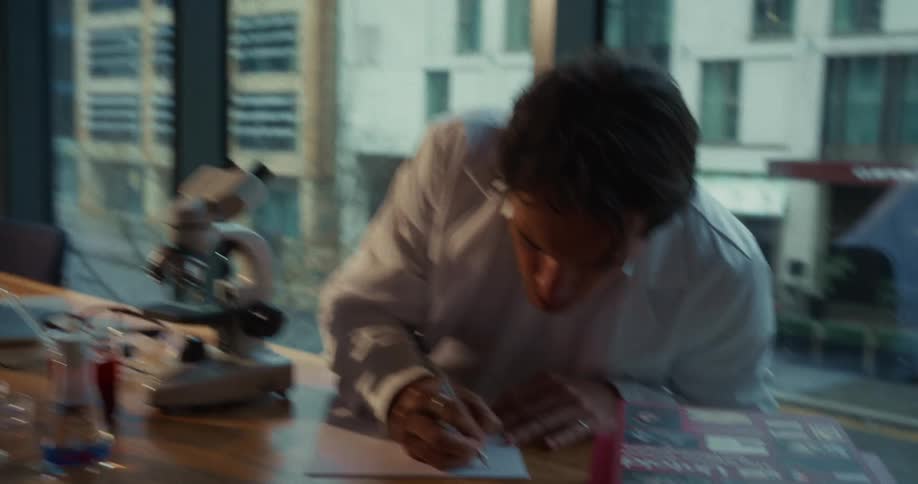 Researcher in a white lab coat observing a sample through a microscope on a bench with jars and a laptop