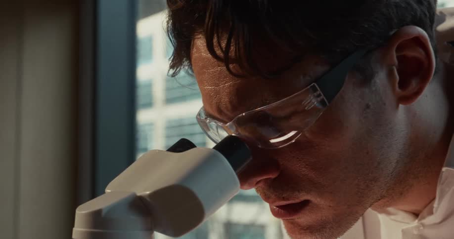 A man wearing a lab coat puts on safety glasses and looks through a microscope