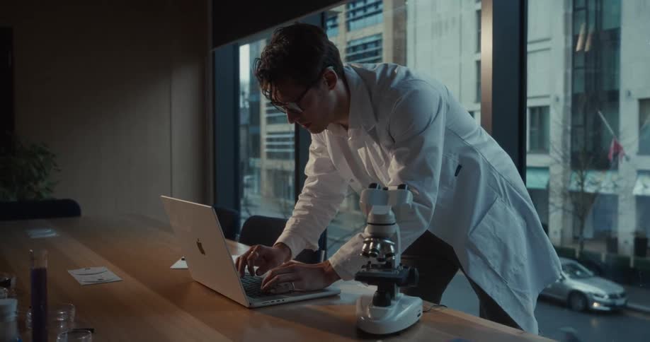 Researcher in a white lab coat types on a laptop beside a microscope on a table near a window with an urban view