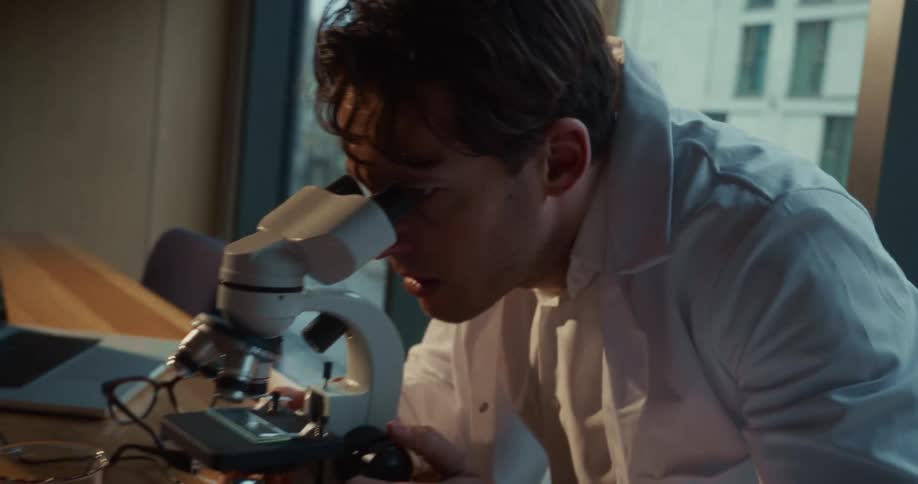 Doctor walks into the room and sits down looking into a microscope and writing notes