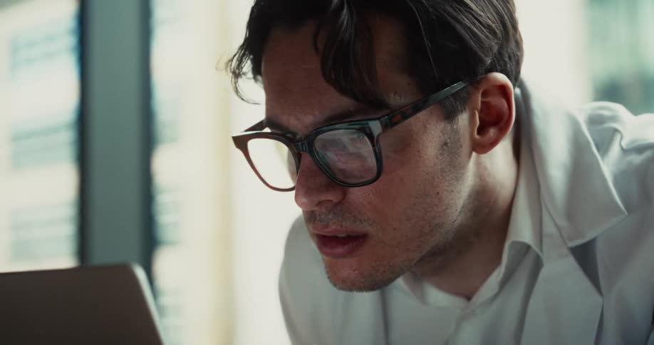 A man wearing glasses and a white lab coat looks at a computer screen in a bright office