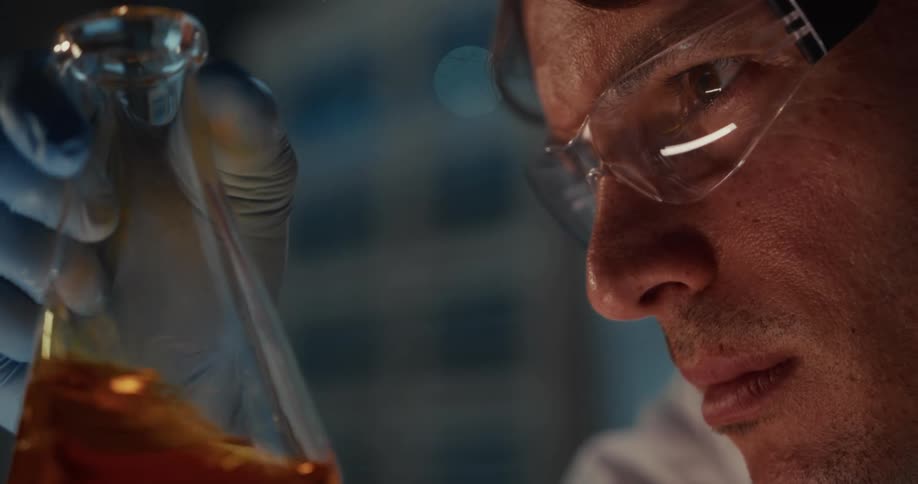 Scientist wearing safety goggles observes a flask containing orange liquid in a laboratory