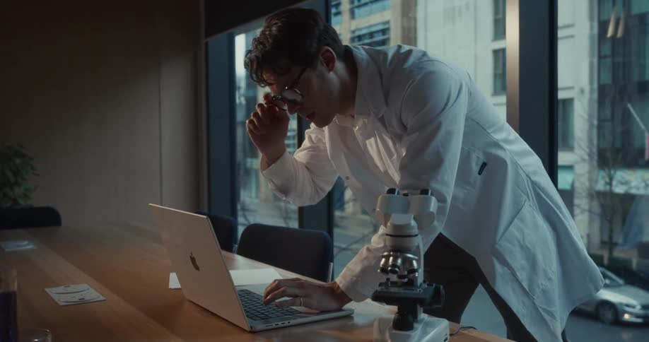 A man wearing a white lab coat and glasses stands at a long wooden table while typing on a silver laptop
