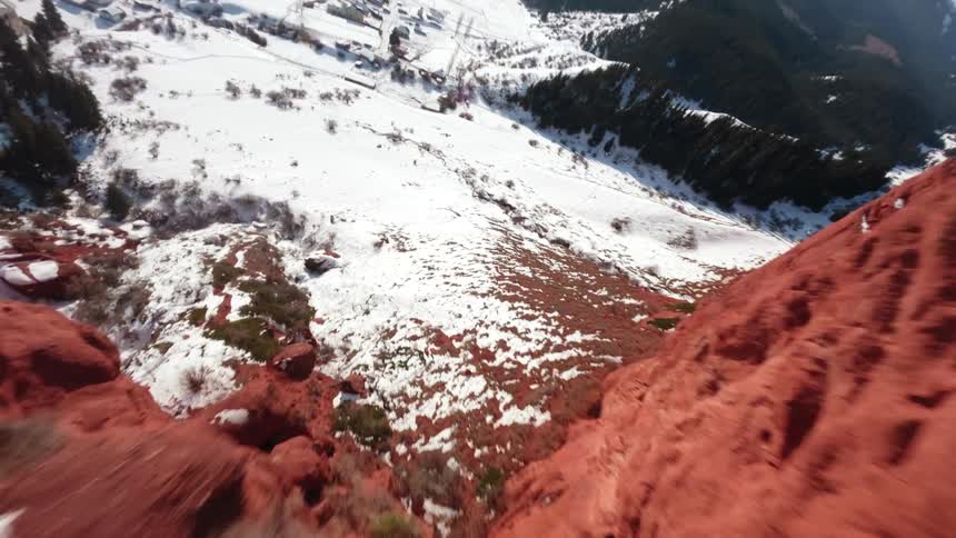 A drone flies rapidly over a narrow red rock ridge lightly dusted with snow before diving down into a wide valley with small houses and dark green trees under a clear blue sky