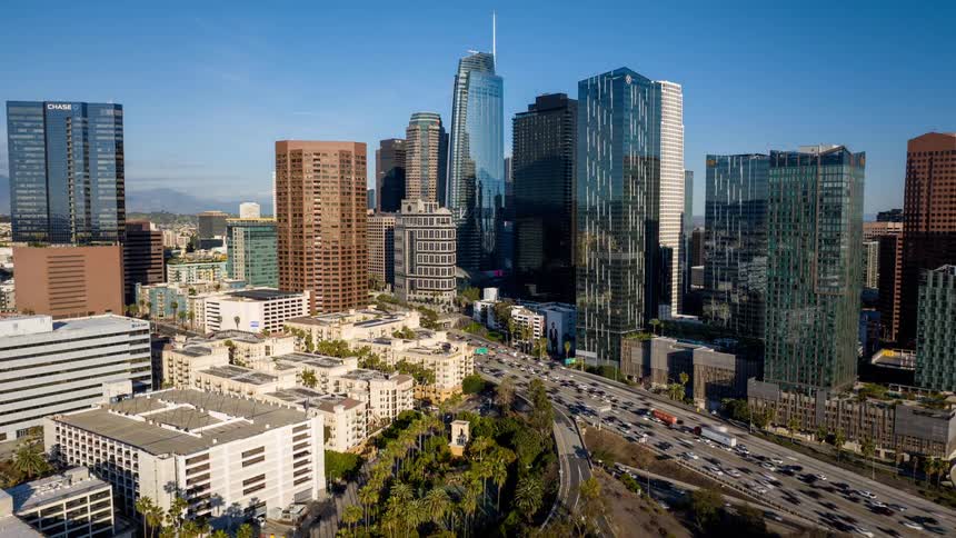 A high view shows many tall glass skyscrapers and office buildings in Downtown Los Angeles under a clear blue sky as many cars move along a wide highway during the day