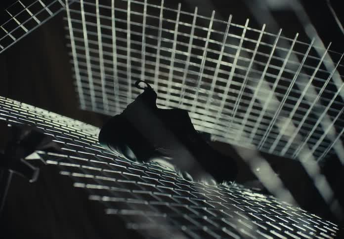 A black athletic shoe sits on a metal wire grid as more metal grids spin and move around it in a dark room with dramatic lighting