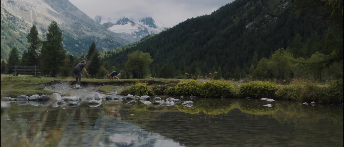 A man and a young boy run along the grassy bank of a shallow stream in a mountainous region the man performs a cartwheel while the child watches from a distance as they explore the scenic landscape